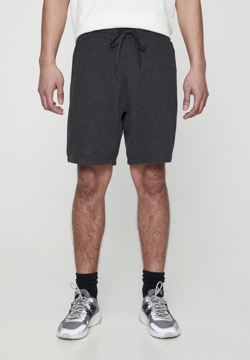 Shorts gris texturés avec une taille élastique et un cordon de serrage. Portés avec des baskets grises et des chaussettes foncées, debout devant un fond uni.