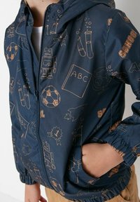 Child wearing navy blue jacket with school-themed tan illustrations including ABC book, robot, soccer ball, rocket, test tubes, and "SCHOOL" text.