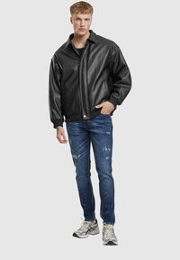 Chaqueta de cuero sintético negra con puños y cuello acanalados, combinada con jeans azules desgastados y zapatillas de running plateadas sobre un fondo simple.