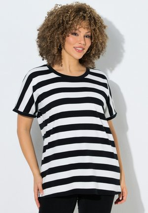 Frau mit lockigem Haar, die ein schwarz-weiß gestreiftes Kurzarmshirt trägt und vor einem einfarbigen hellen Hintergrund lächelt.