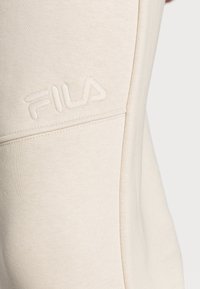 Beige Fila jogger met een zachte textuur, voorzien van een reliëf logo en een aansluitend ontwerp dat de benen vormt. Zijdeteen detail zichtbaar.