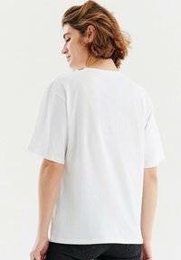 Bílý bavlněný tričko s krátkými rukávy, volný střih a kulatým výstřihem. Hladká textura a žádné viditelné vzory nebo grafické designy.
