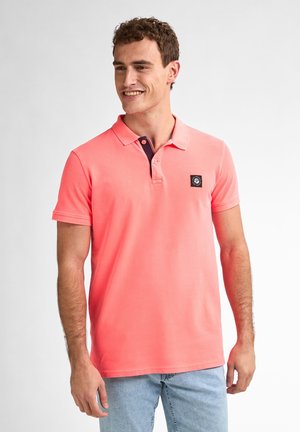 Korallinvärinen polo-paita lyhyillä hihoilla, jossa on tummansininen kaulus ja pieni musta logo-ompelu vasemmassa rinnassa. Silkkisen pehmeä puuvillamateriaali.
