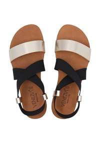 Zwarte en gouden sandalen met een platte zool, uitgerust met een brede metallische band en twee elastische banden die over een bruine voetbed kruisen.