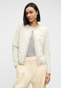 Γυναίκα με κοντά σκούρα μαλλιά που φοράει ανοιχτόχρωμη φλοράλ ζακέτα πάνω από γκρι ριμπ μπλούζα και μπεζ παντελόνι με πιέτες και ψηλή μέση, με τα χέρια στις τσέπες.