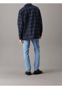 Černá a modrá košile s kostkovaným vzorem ve stylu overshirt s límcem, mírně volný střih. Oblečení doplňují světle modré džínové kalhoty a černé boty.