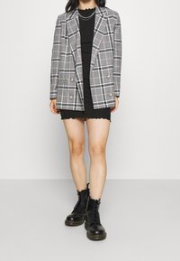 Šedý kostkovaný dvouřadý blazer s růžovými akcenty, kovovými knoflíky a klopami, vrstvený přes černé žebrované šaty s vlněným lemem.