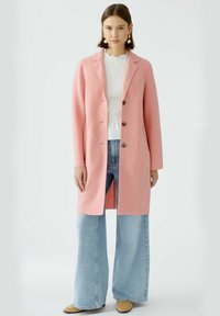Langer rosa Mantel aus strukturiertem Stoff, mit Reverskragen, fünf Knöpfen und seitlichen Schlitzen, kombiniert mit weit geschnittenen blauen Jeans und beigen Schuhen.