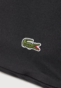 Bolsa textil negra con un logo de cocodrilo verde con detalles rojos, contorneado en bordado blanco. La tela tiene un acabado texturizado.