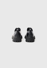 Scarpe in pelle nera con una suola in gomma spessa e rigida e cuciture sottili. Presentano un'etichetta con logo sul retro e un design a punta rotonda.