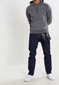 Felpa grigia in pile con tasca frontale, abbinata a jeans blu scuro con cuciture a contrasto e un bandana nera infilata in vita. Sneakers bianche.
