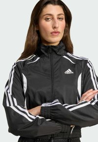 Giacca nera in nylon con zip, strisce bianche sulle maniche, colletto alto e polsini elastici. Presenta il logo Adidas sul petto.