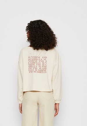 Woman with curly hair wearing a cream long-sleeve top and matching pants, back facing, with "ROXY" and repeated text in red on the top.