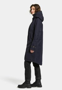 Mörkblå hooded parka med rak skärning, utrustad med dragkedja framtill, sidofickor och knäppningar vid ärmsluten, tillsammans med svarta jeans och stövlar.