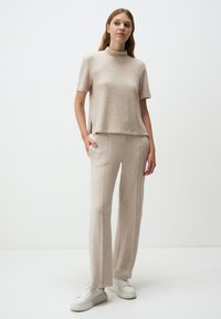 Beige gebreide top met korte mouwen en een opstaande kraag, gecombineerd met bijpassende beige wijde broek. Beiden hebben een zachte textuur. Witte sneakers maken de look compleet.