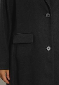 Czarny wełniany płaszcz w środkowej części z dwoma guzikami i kieszenią z klapą, pokazujący dłoń z naturalnymi paznokciami z boku.