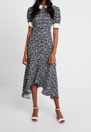 Woman wearing a black and white patterned midi dress with puff sleeves, white cuffs, and a white collar, paired with white ankle-strap heels.