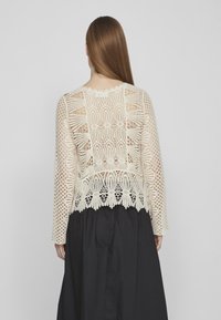 Top in pizzo all'uncinetto color avorio con maniche lunghe, motivo intricato, orlo ondulato e una texture semitrasparente. Indossato su una gonna nera.