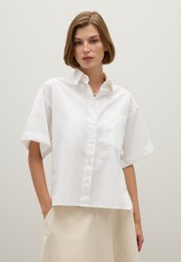 White cotton short-sleeve shirt with a button-down front, left chest pocket, and relaxed fit. Smooth texture, slightly cropped design.
