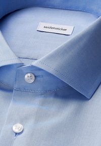 Camicia azzurra chiara con una texture di tessuto liscia, colletto appuntito e un'etichetta logo bianca. Presenta bottoni bianchi sul colletto.