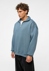 Lichtblauwe, oversized sweatshirt met een ritskraag, lange mouwen en geribbelde manchetten. Zachte textuur, minimale stikseldetails. Draag het met donkere broeken.