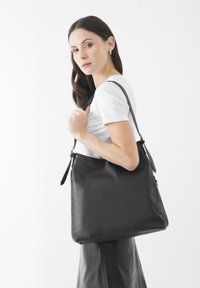 Zwarte leren tote bag met een gladde textuur, voorzien van een enkele verstelbare schouderband en een subtiele vouw aan de bovenkant.