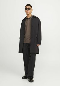 Čierny dlhý kabát cez hnedé polo tričko, skombinovaný s tmavými nohavicami a čiernymi topánkami. Doplnky zahŕňajú tmavé slnečné okuliare. Hladká, štruktúrovaná látka.