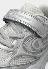 Λευκό αθλητικό παπούτσι με ανώτερο μέρος από διχτυωτό υλικό, ασημένιες λεπτομέρειες και ιμάντα με βέλκρο. Εμφανής υφή στα πλαϊνά και επένδυση στο κολάρο.