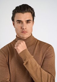 Maglione a collo alto marrone realizzato in morbido tessuto lavorato a maglia; design aderente con polsini e scollo a costine; texture liscia senza motivi.