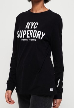 Osoba ubrana w czarną koszulę z długim rękawem z napisem „NYC SUPERDRY” oraz współrzędnymi wydrukowanymi na piersi białą czcionką.