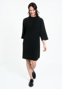 Černé šaty do kolen s nadměrně velkými rukávy, vyrobené z hladké, měkké látky. Mají kulatý výstřih a minimální detaily švů.