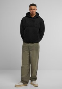 Schwarzer Pullover-Hoodie mit frontaler Tasche, ausgestattet mit erhabener "MARKET"-Aufschrift. Kombiniert mit olivgrünen weiten Hosen und sneakers mit beigen Akzenten.