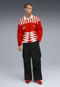 Crveno-bijeli prugasti pulover s ovratnikom, koji sadrži Puma logo i grafičke detalje, u kombinaciji s labavim crnim cargo hlačama i crvenim cipelama.