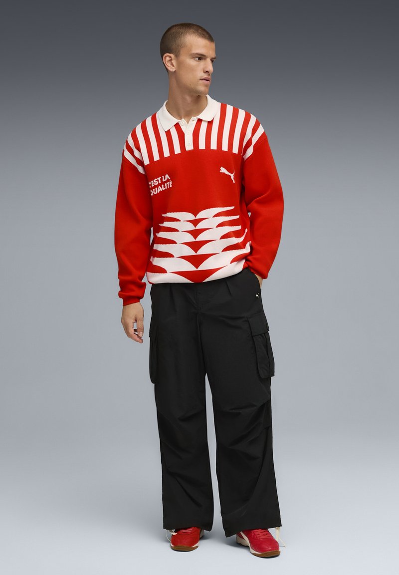Crveno-bijeli prugasti pulover s ovratnikom, koji sadrži Puma logo i grafičke detalje, u kombinaciji s labavim crnim cargo hlačama i crvenim cipelama.