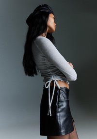 Sivý dlhý rukáv top s nariasenými bočnými viazaniami, skombinovaný s čiernou koženou mini sukňou so striebornými nitmi. Pozadie je tlmenej sivej farby.