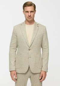 Férfi áll szemben, bézs textúrájú zakóval és hozzáillő nadrággal, alatta világosbézs pólóval.