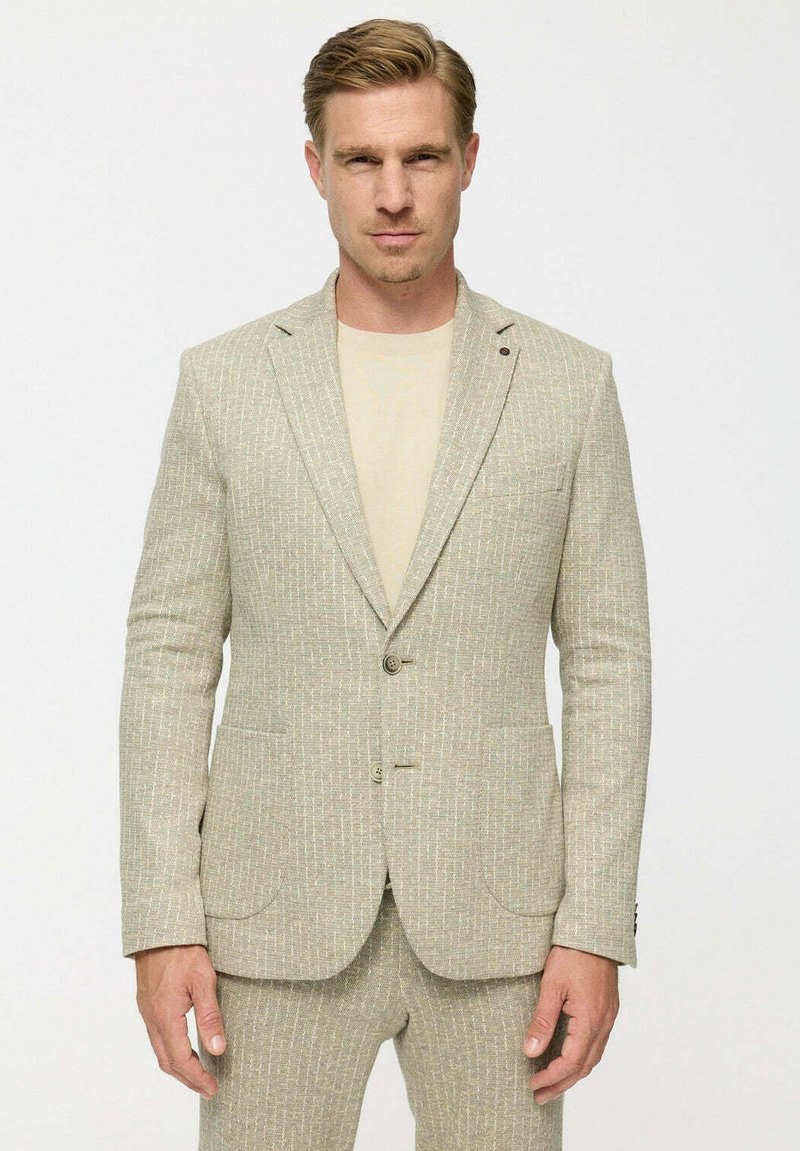 Férfi áll szemben, bézs textúrájú zakóval és hozzáillő nadrággal, alatta világosbézs pólóval.