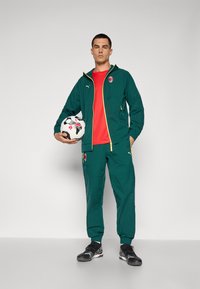 Grøn tracksuit med gule detaljer, rød skjorte og sorte sko. Holder en fodbold med et holdlogo på toppen.