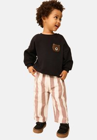 Felpa nera con toppa a forma di orso, pantaloni a gamba larga beige con righe rosa, sneakers nere con chiusure a strappo. Bambino in piedi che sorride.