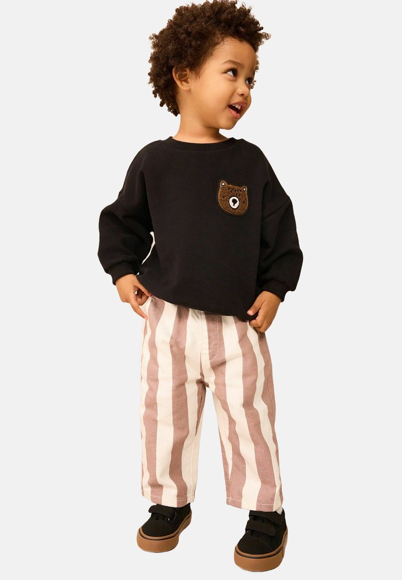Felpa nera con toppa a forma di orso, pantaloni a gamba larga beige con righe rosa, sneakers nere con chiusure a strappo. Bambino in piedi che sorride.