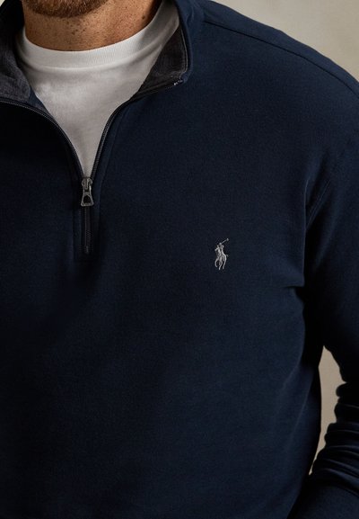 Μπλε ναυτικό πουλόβερ με γιακά μισού φερμουάρ και γκρι εσωτερική επένδυση. Διαθέτει ένα μικρό κεντημένο λογότυπο στο στήθος. Απαλή, υφή σαν φλις.