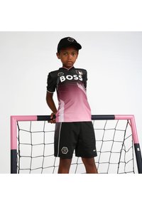 Un garçon portant un maillot de soccer noir et rose dégradé avec le logo "BOSS", associé à un short noir et une casquette noire, se tenant à côté d'un poteau de but rose.