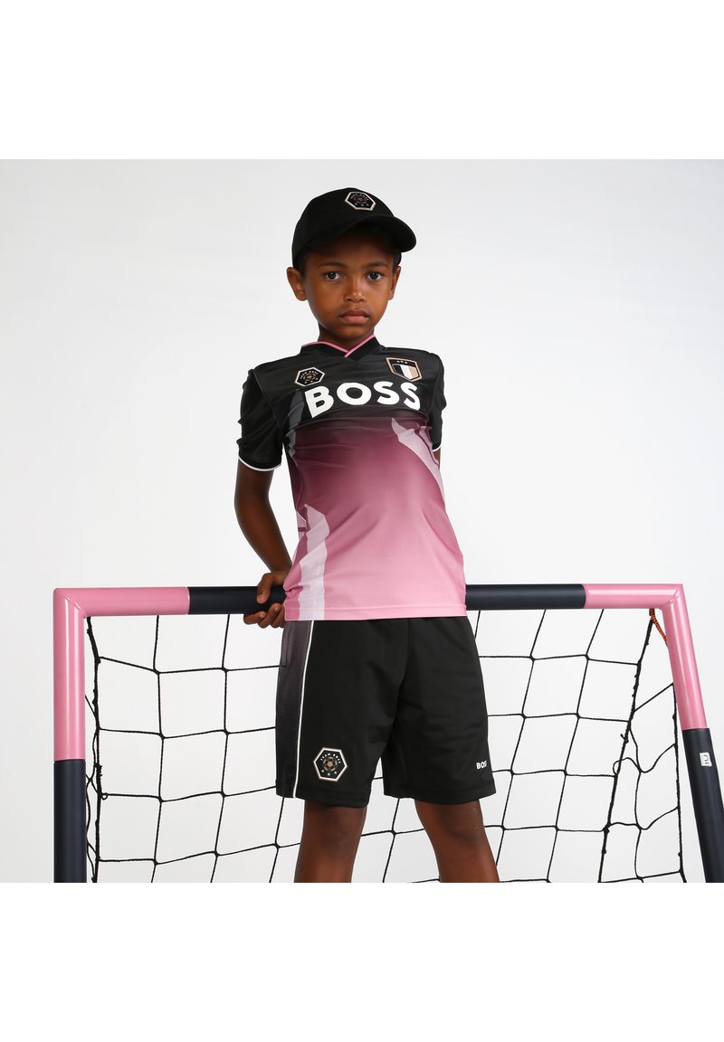 Un garçon portant un maillot de soccer noir et rose dégradé avec le logo "BOSS", associé à un short noir et une casquette noire, se tenant à côté d'un poteau de but rose.