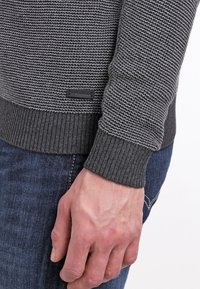 Maglione grigio a maglia con un motivo a righe testurizzato, con polsini e orlo a costine, e una piccola etichetta nera in basso. Mani che tengono il maglione.