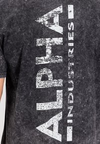 Crna majica s acid-wash efektom, sa velikim vertikalnim bijelim istrošenim natpisom "ALPHA INDUSTRIES" sprijeda, koju nosi osoba vidljiva s lijeve strane.
