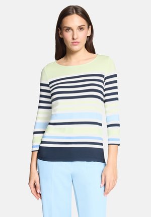 Femme portant un haut à manches longues rayé en bleu marine, vert clair, blanc et bleu, associé à un pantalon bleu clair, debout devant un fond uni.