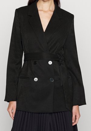 Frau trägt ein tailliertes, schwarz zweireihiges Sakko mit Taillengürtel und einen plissierten schwarzen Rock, die Ärmel sind lang und gerade.