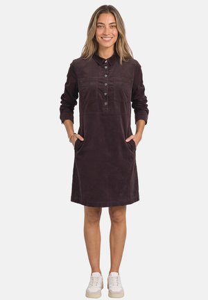 Femme souriante portant une robe boutonnée à manches longues de couleur violet foncé avec des poches et des baskets blanches, debout devant un fond blanc uni.
