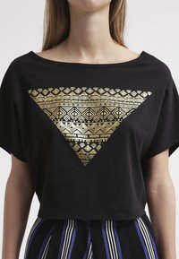 Camiseta negra de corte cropped con un patrón geométrico dorado en forma de triángulo en el frente. Manga corta y un diseño holgado.