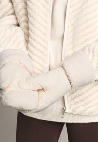 Krem-bijela umjetna krzna jakna s vertikalnim prugama, koja ima mekan, teksturiran završetak i široke flis manžete, uparen s off-white puf rukavicama.